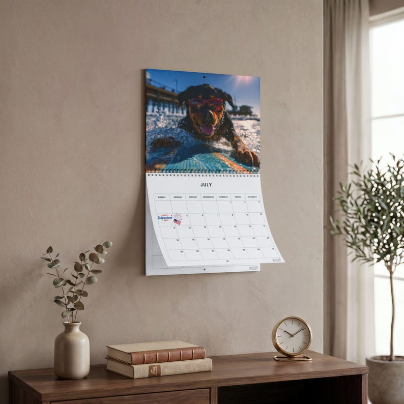 K&ouml;nnte beinhalten: Ein Wandkalender f&uuml;r Juli mit einem Foto eines schwarzen Hundes, der eine rote Sonnenbrille tr&auml;gt und auf einem blauen Surfbrett surft. Das Kalenderraster enth&auml;lt den Text July und ein kleines Symbol einer amerikanischen Flagge am vierten Tag.