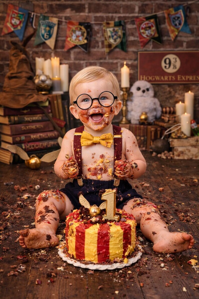 May include: A baby covered in cake, wearing round glasses, a yellow bow tie, and suspenders, celebrating a first birthday. A cake with red and yellow stripes and a golden "1" sits in front of the baby. The background includes Harry Potter-themed decorations.