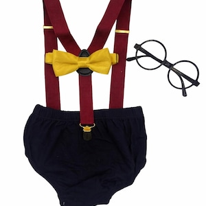 May include: A baby outfit set with a navy blue nappy cover, burgundy braces with gold details, and a yellow bow tie. Includes round black glasses. Ideal for a themed photoshoot or special event.