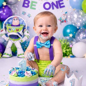 May include: A smiling baby with frosting on their face and hands, wearing a blue bow tie and suspenders, celebrates their first birthday. A colorful cake with a number one candle and a crown are in front of the baby. Balloons and a banner that says "INFINITY AND BE ONE" are in the background.