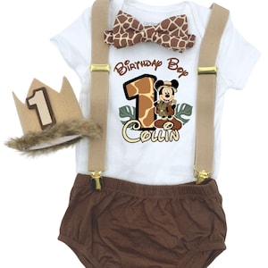 May include: A white bodysuit with a Mickey Mouse graphic, brown shorts, suspenders, and a bow tie with a giraffe print. The outfit includes a crown with the number "1" on it. The text "Birthday Boy" and a name are printed on the bodysuit.