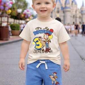 May include: A young child wearing a cream-colored t-shirt and blue shorts. The shirt features a graphic of Woody from Toy Story with the text "It's My Birthday" and the number 3. The shorts have a matching Woody graphic.