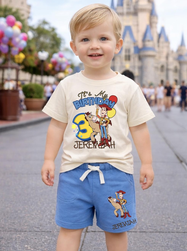 May include: A young child wearing a cream-colored t-shirt and blue shorts. The shirt features a graphic of Woody from Toy Story with the text "It's My Birthday" and the number 3. The shorts have a matching Woody graphic.