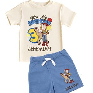 May include: A two-piece children's outfit featuring a cream-colored t-shirt and blue shorts. The shirt has the text "It's My Birthday" and a graphic of a cartoon cowboy. The shorts also have a matching graphic and the name "Jeremiah".