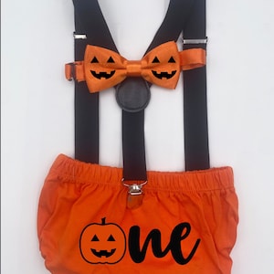 May include: An orange Halloween outfit for a one-year-old, featuring orange shorts with a pumpkin and the word "one", black suspenders, and a pumpkin bow tie. The bow tie and shorts have jack-o'-lantern faces.