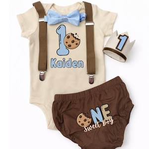 May include: A beige baby outfit set with a blue bow tie, suspenders, and a crown. The onesie has the number 1 and a cookie graphic, with the name "Kaidan". Brown diaper cover with "ONE Sweet Boy" text and cookie graphic.