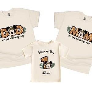 May include: Three cream-colored t-shirts with a jungle theme. The shirts feature a cartoon Mickey Mouse wearing a safari hat and holding a magnifying glass. The text on the shirts reads "Dad of the birthday boy", "Mom of the birthday boy", and "Birthday Boy" with the number "1" and the name "Wilson".