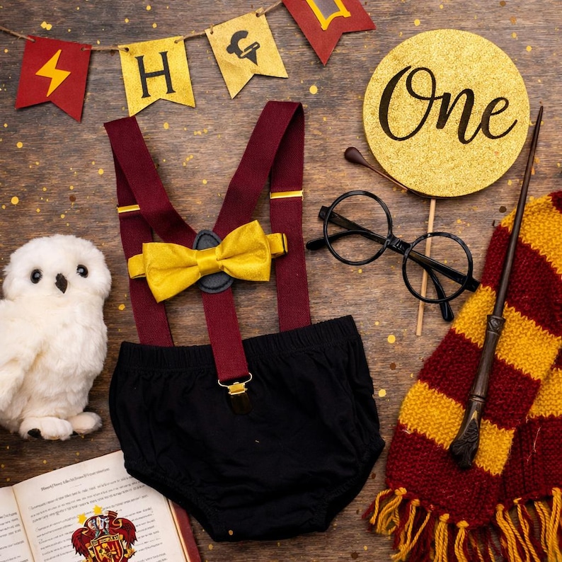 May include: A themed first birthday set with a black nappy cover, burgundy braces, and a yellow bow tie. Accessories include round glasses, a wand, a scarf, and a plush owl. A banner spells out "H" and a gold circle says "One".
