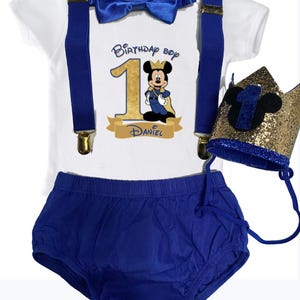 May include: A white onesie with blue suspenders and a blue bow tie. The onesie has a gold number one with a Mickey Mouse character wearing a crown and a cape. The text "Birthday Boy" and "Daniel" are printed on the onesie. A blue and gold crown with a Mickey Mouse ear is included.