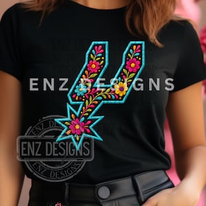 Puede incluir: Camiseta negra con un diseño floral colorido en forma de espuela. El diseño incluye flores rosas, amarillas y azules con un contorno turquesa. El texto "ENZ DESIGNS" está impreso en la camiseta.