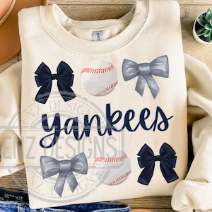 May include: Cream-colored sweatshirt featuring the word "Yankees" in navy blue script, along with baseballs and bows. The bows are navy and gray. The baseballs have red stitching. The sweatshirt has a crew neck.