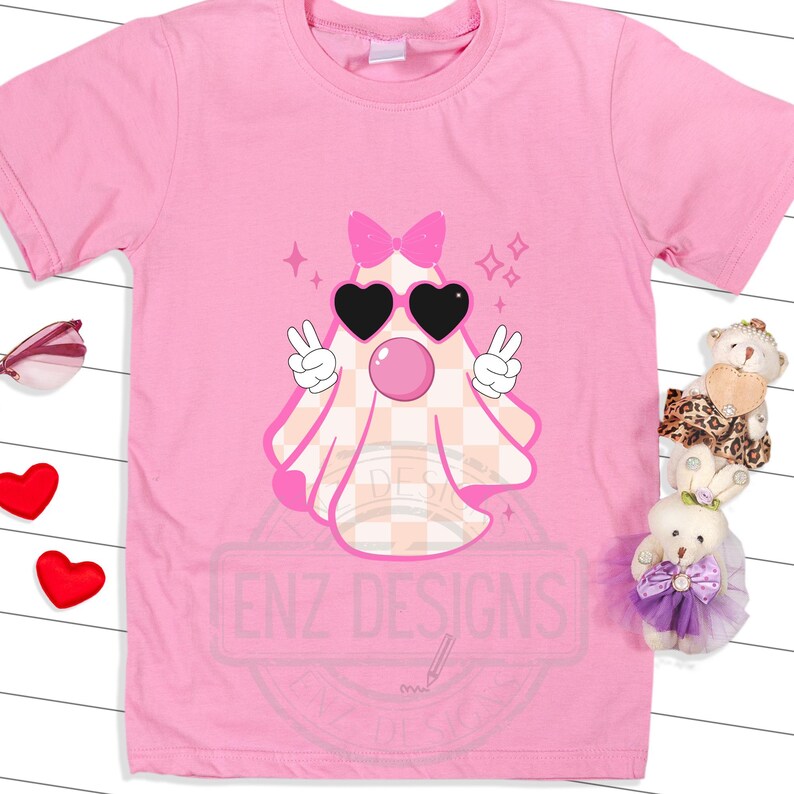 Op de afbeelding: Roze T-shirt met een roze en wit geruite geest die een zonnebril en een roze strik draagt. De geest heeft een roze bel die uit zijn mond komt. De tekst "ENZ DESIGNS" is op het T-shirt gedrukt.