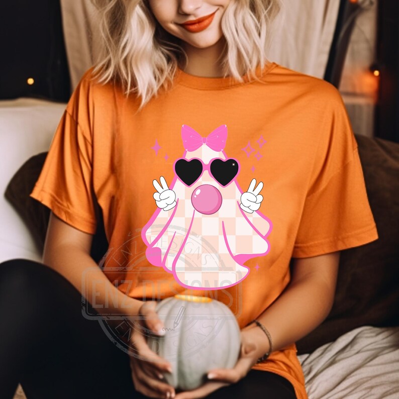 Op de afbeelding: Oranje T-shirt met een roze en wit geruite geest die een zonnebril en een roze strik draagt. De geest maakt een peace-teken met beide handen. De tekst "BOO" staat boven de geest.