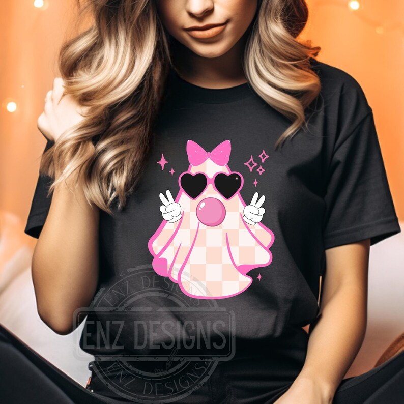 Op de afbeelding: Een zwart T-shirt met een roze en wit geruite geest die hartvormige zonnebrillen en een roze strik draagt. De geest maakt een peace-teken met beide handen. De tekst "ENZ DESIGNS" is zichtbaar op het shirt.