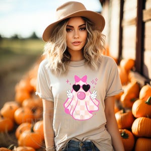 Op de afbeelding: Een vrouw draagt een beige t-shirt met een roze spook dat een zonnebril en een strik draagt. Het spook heeft een geruit patroon en maakt peace-tekens. De vrouw staat voor een stapel pompoenen.
