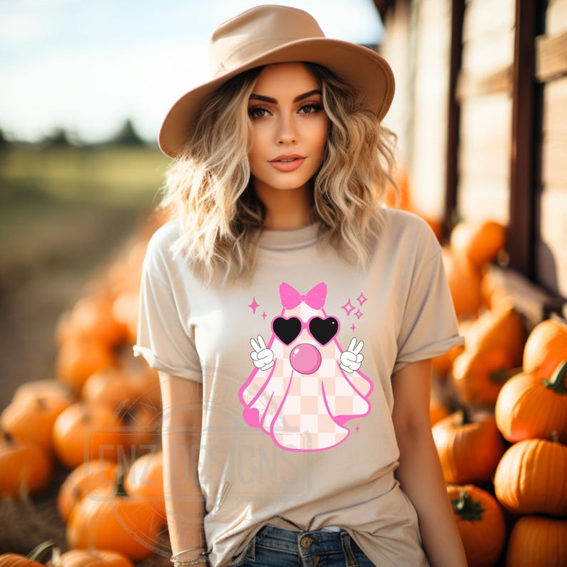 Op de afbeelding: Een vrouw draagt een beige t-shirt met een roze spook dat een zonnebril en een strik draagt. Het spook heeft een geruit patroon en maakt peace-tekens. De vrouw staat voor een stapel pompoenen.