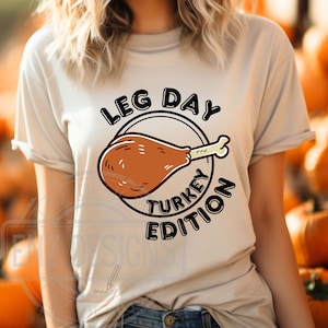Könnte beinhalten: Ein beiges T-Shirt mit einem schwarzen Kreis-Motiv, auf dem "Leg Day Turkey Edition" steht und ein Cartoon-Bild eines Truthahnbeins zu sehen ist.