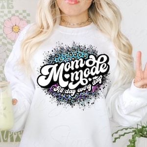 May include: White sweatshirt with a graphic print of a blue, pink, and black splatter design with leopard print accents. The graphic says "Mom Mode All day every day" in white text with a heart in the middle of "Mom" and a black outline.