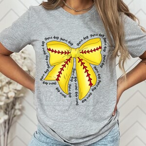 K&ouml;nnte beinhalten: Graues T-Shirt mit einem gelben und roten Softball-Schleifendesign. Die Schleife ist von dem Text "game day" umgeben, der mehrmals wiederholt wird.