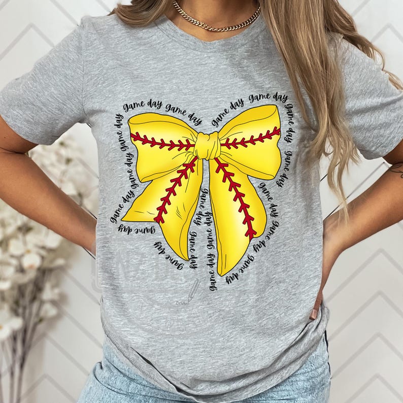 K&ouml;nnte beinhalten: Graues T-Shirt mit einem gelben und roten Softball-Schleifendesign. Die Schleife ist von dem Text "game day" umgeben, der mehrmals wiederholt wird.