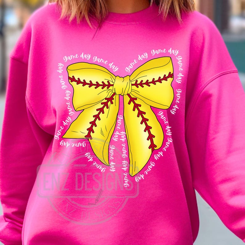 K&ouml;nnte beinhalten: Ein rosa Sweatshirt mit einem gelben und roten Softball-Schleifendesign. Die Schleife ist von dem Text "game day" umgeben, der mehrmals wiederholt wird.