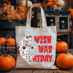 K&ouml;nnte beinhalten: Eine wei&szlig;e Canvas-Tragetasche mit einer roten und wei&szlig;en Grafik eines Hockey-Masks mit Blutspritzern und dem Text "I WISH IT WAS FRIDAY". Die Tasche liegt auf einer Holzoberfl&auml;che mit K&uuml;rbissen im Hintergrund.