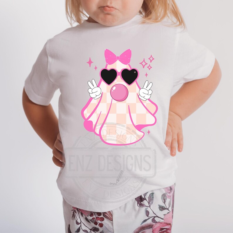 Op de afbeelding: Een wit T-shirt met een roze en wit geruite geest die een roze zonnebril en een roze strik draagt. De geest maakt een peace-teken met beide handen en heeft een roze bel die uit zijn mond komt. Het shirt heeft de tekst "ENZ DESIGNS" onderaan.