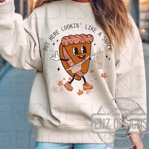 Puede incluir: Una sudadera blanca con un personaje de pastel de dibujos animados marrón y naranja que sostiene una bebida. El personaje del pastel lleva una riñonera marrón y tiene el texto "Out here lookin' like a snack" a su alrededor.