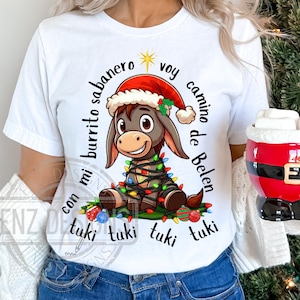 Könnte beinhalten: Weißes T-Shirt mit einem Cartoon-Esel, der einen Weihnachtsmann-Hut und Weihnachtslichter trägt. Der Text auf dem Shirt lautet "con mi burrito sabanero voy camino de Belen tuki tuki tuki tuki"
