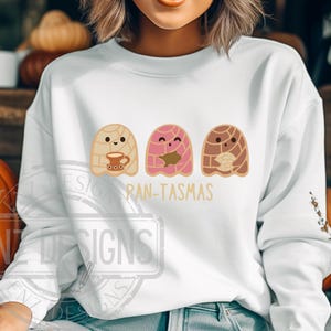 May include: White sweatshirt with a graphic of three cartoon ghosts shaped like pan dulce, a traditional Mexican sweet bread. The ghosts are holding a cup of coffee, a piece of pan dulce, and a piece of pan dulce. The text "Pan-Tasmas" is printed below the ghosts.