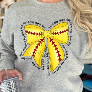 K&ouml;nnte beinhalten: Ein graues Sweatshirt mit einem gelben und roten Softball-Schleifendesign. Die Schleife ist von dem Text "game day" umgeben, der mehrmals wiederholt wird.