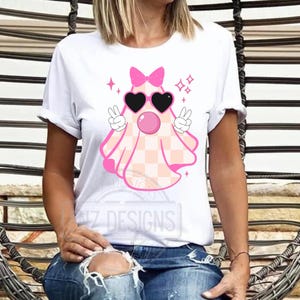 Op de afbeelding: Wit T-shirt met een roze spook dat een zonnebril en een strik draagt. Het spook heeft een geruit patroon en steekt vredestekens omhoog. De tekst "NZ DESIGNS" is zichtbaar op het shirt.