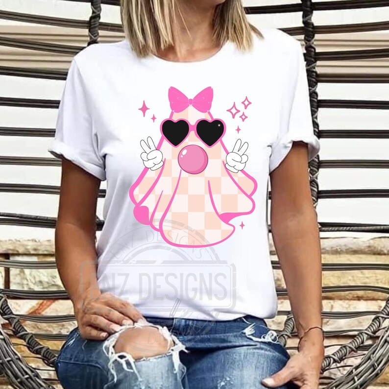 Op de afbeelding: Wit T-shirt met een roze spook dat een zonnebril en een strik draagt. Het spook heeft een geruit patroon en steekt vredestekens omhoog. De tekst "NZ DESIGNS" is zichtbaar op het shirt.