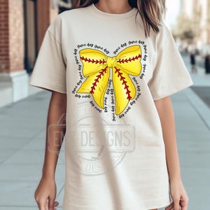 K&ouml;nnte beinhalten: Ein beiges T-Shirt mit einem gelben und roten Softball-Schleifendesign. Die Schleife ist von dem Text "game day" umgeben, der mehrmals wiederholt wird.