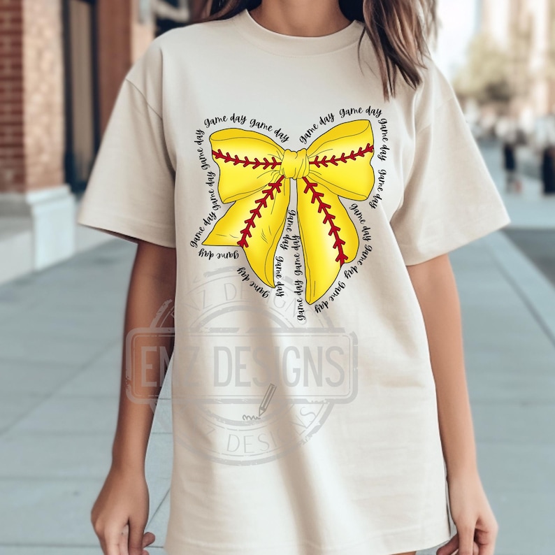K&ouml;nnte beinhalten: Ein beiges T-Shirt mit einem gelben und roten Softball-Schleifendesign. Die Schleife ist von dem Text "game day" umgeben, der mehrmals wiederholt wird.
