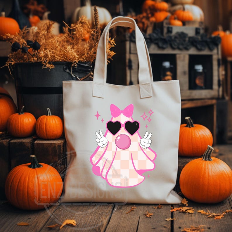 Op de afbeelding: Een witte canvas tote bag met een roze en wit geruite geest die een zonnebril en een roze strik draagt. De geest blaast een bel en heeft vredestekens op zijn handen. De tas ligt op een houten oppervlak met pompoenen op de achtergrond.