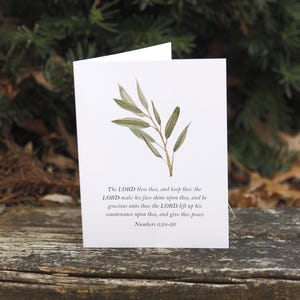 May include: A white greeting card with a watercolour illustration of an olive branch. The card features the text of Numbers 6:24-26. The card is set against a blurred background of green foliage and a wooden surface.