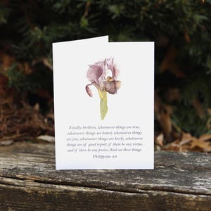 May include: A white greeting card featuring a watercolor painting of a purple iris flower. The card includes text from Philippians 4:8. The card is displayed on a weathered wooden surface with a blurred green background.