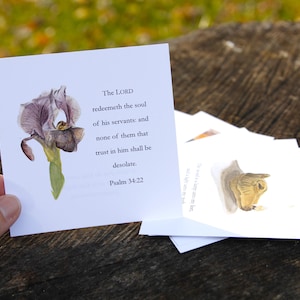 May include: A collection of white square cards featuring watercolour illustrations. One card shows a purple iris flower and text from Psalm 34:22. The cards are on a wooden surface.