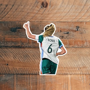 Könnte beinhalten: Ein Aufkleber mit einer Fußballspielerin in einem weißen, türkisblauen und blauen Trikot mit dem Namen "SONIS" und der Nummer "6" auf dem Rücken. Die Spielerin hebt den Daumen, mit blonden Haaren und grünen Shorts. Der Hintergrund ist ein Holzmaserungsmuster.