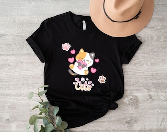 Może przedstawiać: Czarny T-shirt z uroczym kreskówkowym kotem trzymającym serce i otoczonym różowymi sercami i śladami łap. Tekst "Cute" jest nadrukowany pod kotem.