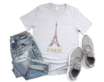 May include: A light gray t-shirt featuring a graphic of the Eiffel Tower and the word "PARIS" in gold. The shirt is paired with blue jeans and gray athletic shoes, creating a casual outfit.