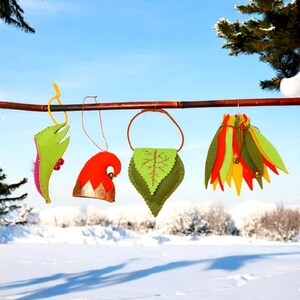 Puede incluir: Cuatro adornos de fieltro coloridos cuelgan de una rama contra un paisaje nevado y un cielo azul. Los adornos incluyen una hoja verde, un gorro de elfo rojo, una bolsa en forma de hoja verde y un adorno en capas.