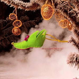 May include: A festive Christmas decoration featuring a green elf shoe with a pink lining and red embellishments. The ornament is suspended by a gold cord, with a backdrop of frosted pine and shimmering golden baubles.