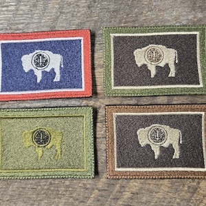 Op de afbeelding: Vier rechthoekige patches met een bisonontwerp. Elke patch heeft een bison-silhouet met een cirkelvormig embleem, omlijst door een rand. De patches hebben verschillende kleurenschema's: rood, groen, bruin en olijf.