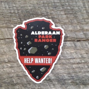 May include: A black and red sticker shaped like a park ranger badge with the text "Alderaan Park Ranger" and "Help Wanted!"  The sticker has a space background with gray asteroids.