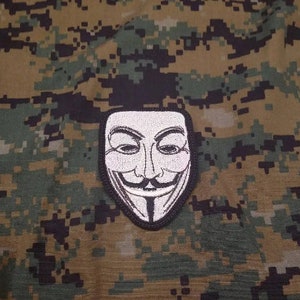 May include: A white embroidered patch of the Anonymous mask on a camouflage background.