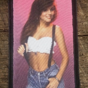 May include: A woman with long brown hair is wearing a white crop top and blue denim overalls. She is standing in front of a pink background.
