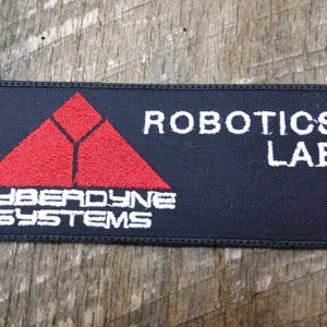 Może przedstawiać: Czarna naszywka haftowana z czerwonym trójkątem i białym tekstem, który brzmi "Cyberdyne Systems" i "Robotics Lab".