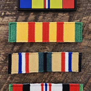 May include: Four rectangular military-style ribbon bars. The top bar is a smooth, black-framed piece with blue, yellow, white, and red segments. The three lower bars are embroidered fabric patches featuring vertical stripes in green, yellow, red, tan, black, white, and blue.